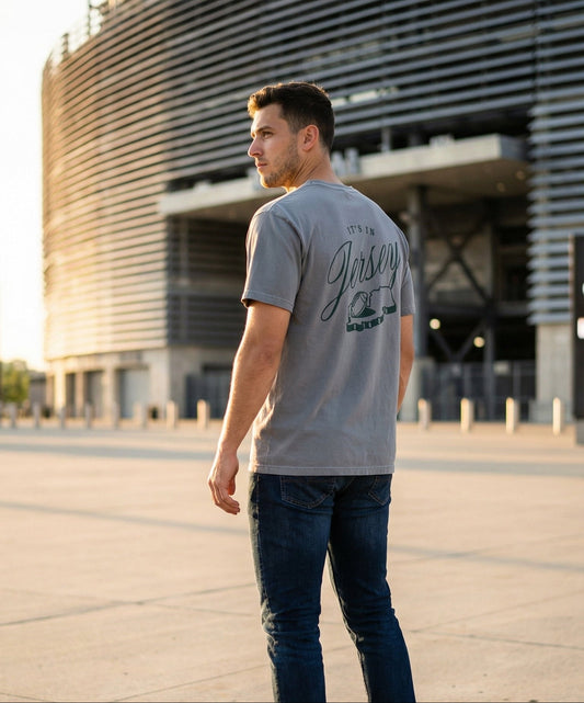 Man wearing a gray t-shirt with It's In Jersey type and the NJ state outline with a football standing in front of a stadium.