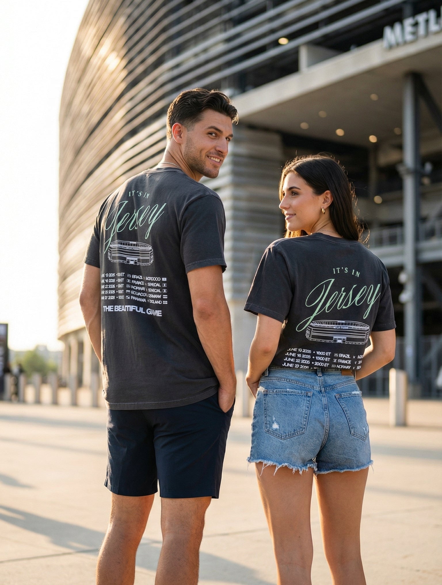 Two people wearing matching grey black t-shirts with It's In Jersey and game info standing in front a stadium.
