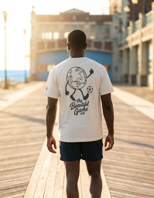 Man walking on the boardwalk wearing a t-shirt that has a large globe playing soccer graphic below that it reads The Beautiful Game 2026