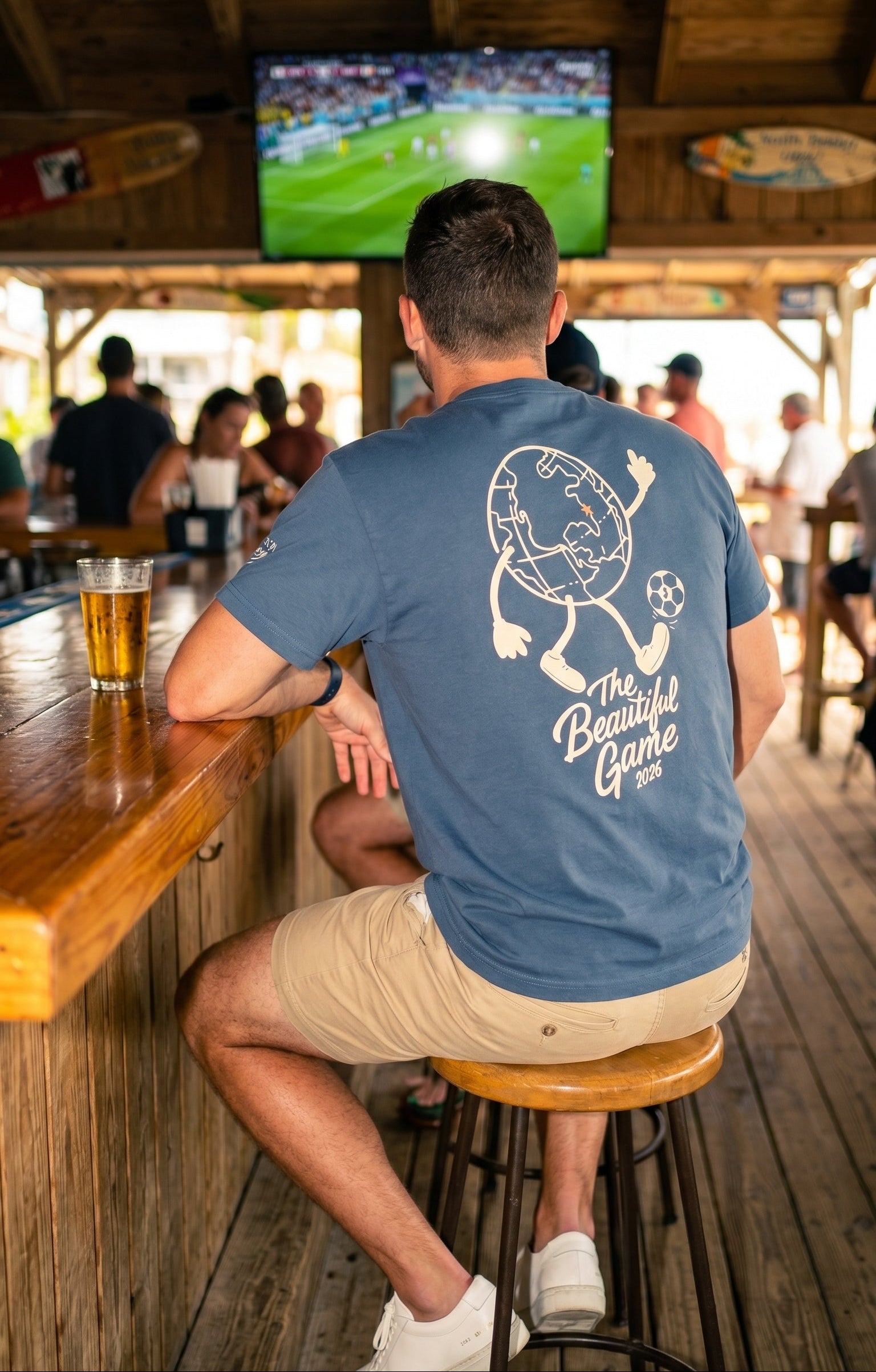 Man sitting at a beach bar wearing a Vintage Navy Tshirt with a large globe kicking a soccer ball and below it ti say The Beautiful Game 2026