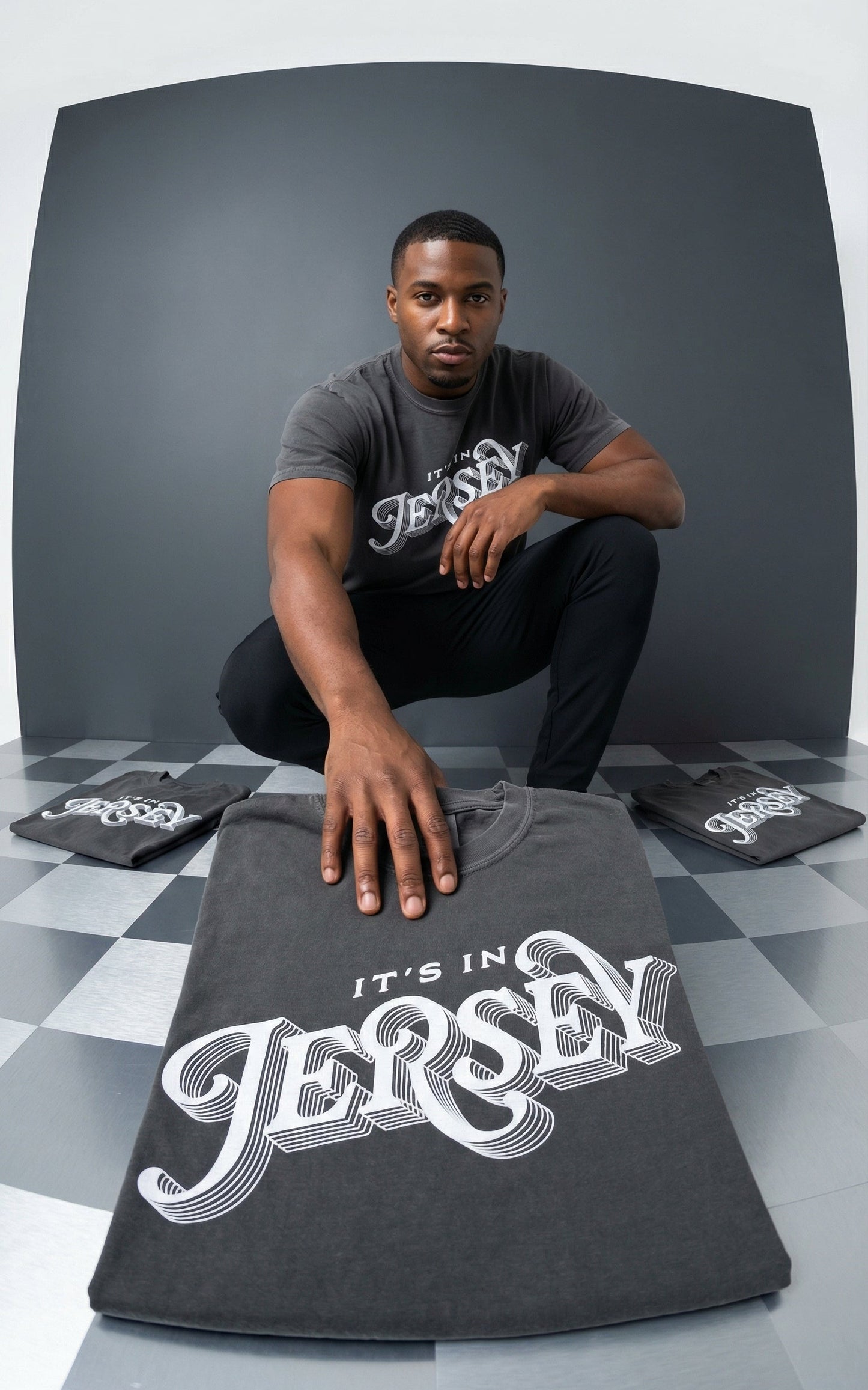 Man holding a t-shirt with 'It's in Jersey' text, surrounded by more t-shirts on a checkered floor.