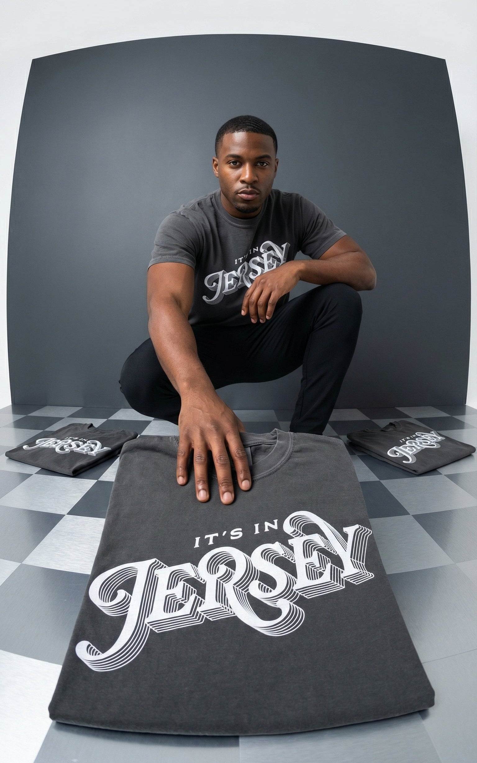 Man holding a t-shirt with 'It's in Jersey' text, surrounded by more t-shirts on a checkered floor.