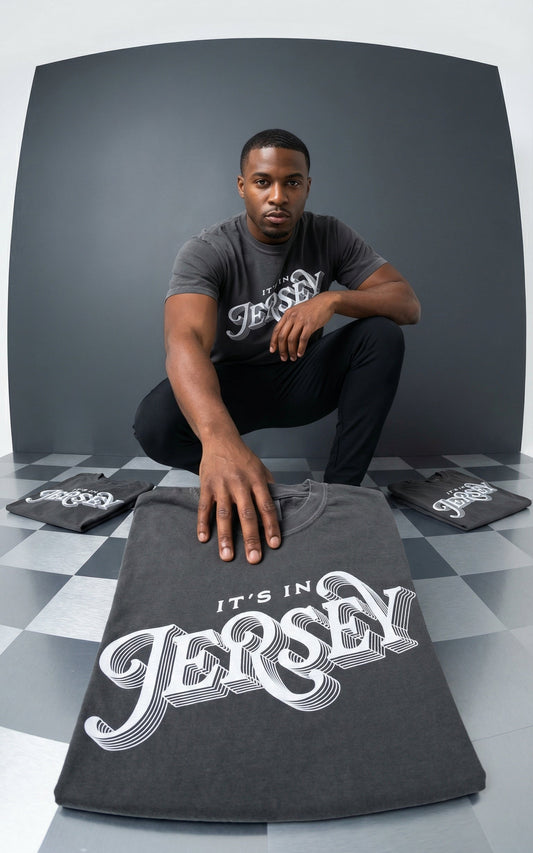 Man holding a t-shirt with 'It's in Jersey' text, surrounded by more t-shirts on a checkered floor.