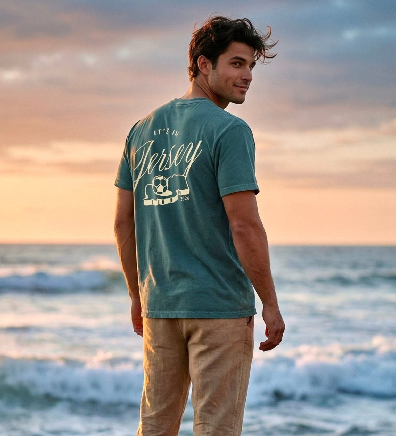 Man standing on rocks by the ocean at sunset wearing a green t-shirt with It's In Jersey text and the state outline with a soccer ball