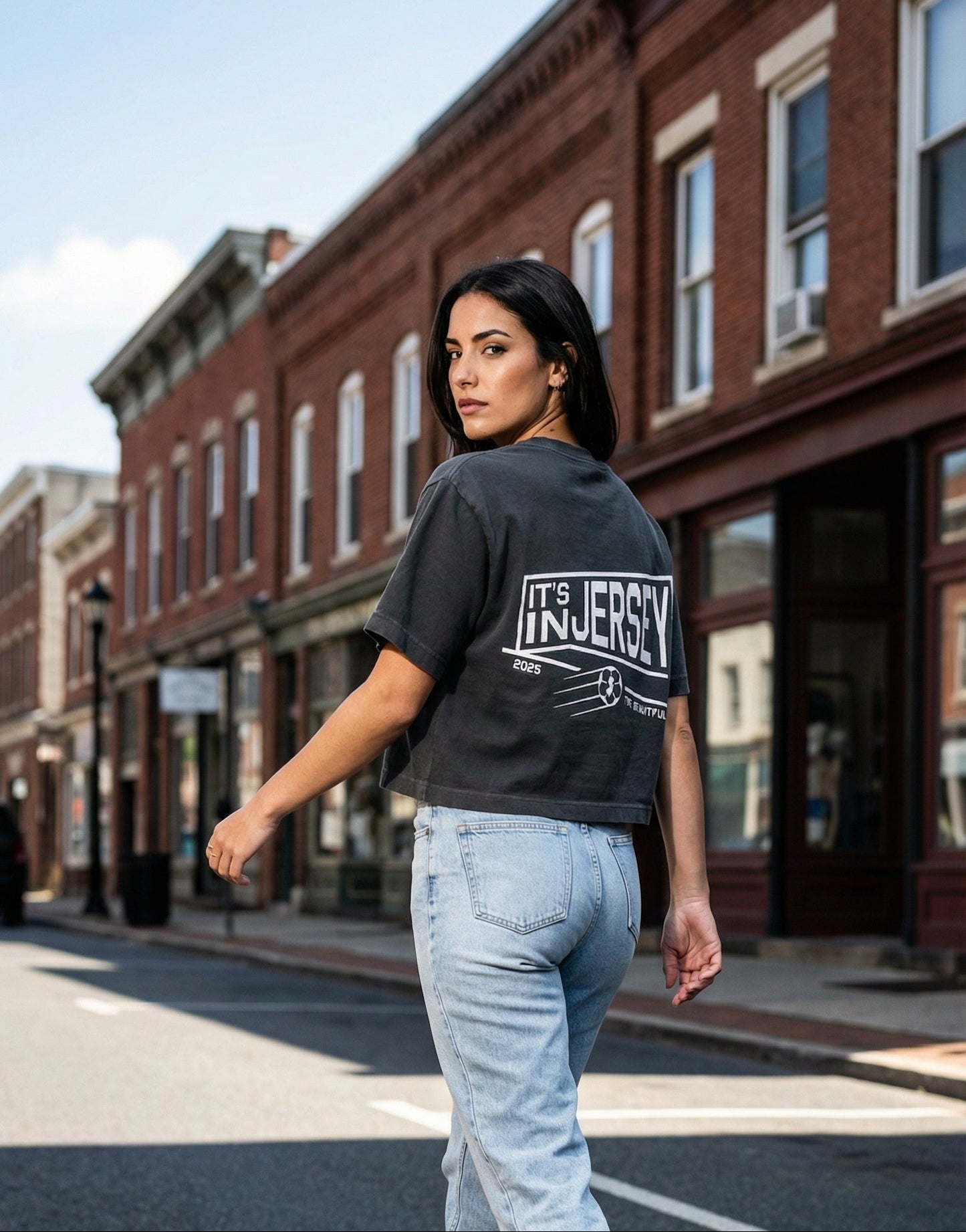 Woman walking on a street wearing a black t-shirt with 'It's In Jersey' text  on the back and light blue jeans.