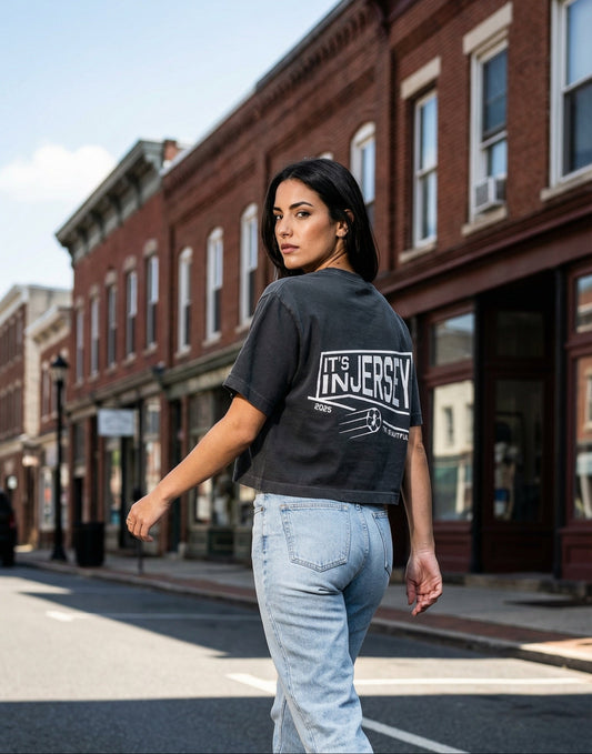 Woman walking on a street wearing a black t-shirt with 'It's In Jersey' text  on the back and light blue jeans.
